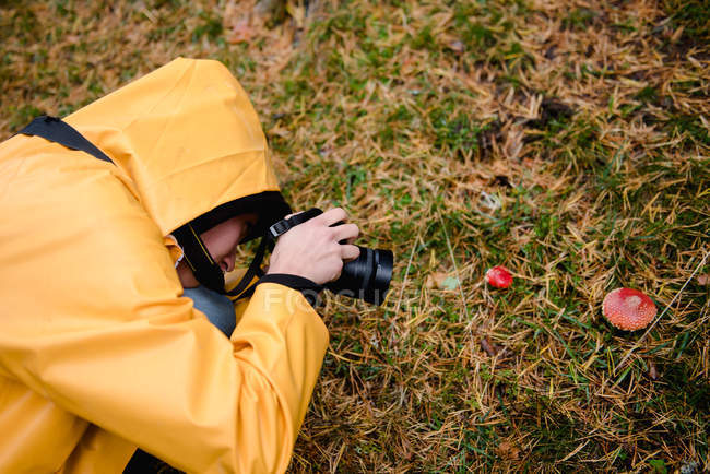 From above of woman in yellow raincoat with hood taking photo of red amanita in grass in forest — Stock Photo