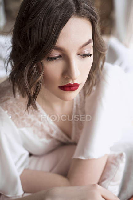Mulher linda com lábios vermelhos em vestido branco olhando para longe enquanto sentado no chão ao lado do sofá — Fotografia de Stock