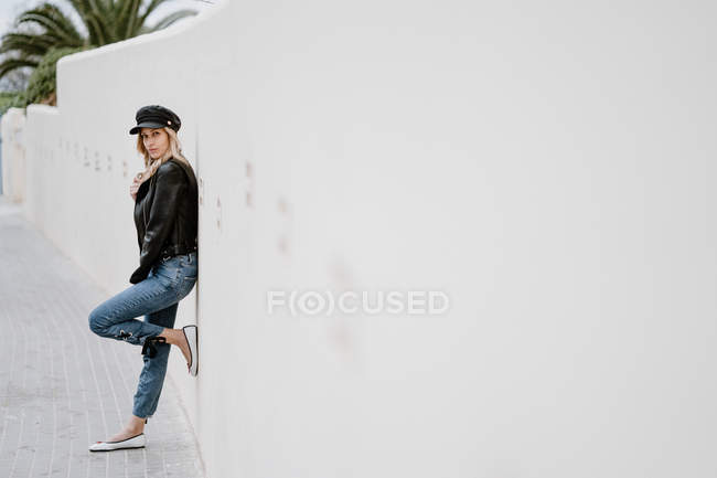 Side view of trendy long haired blonde in black cap and jacket looking at camera and leaning on wall with leg raised — Stock Photo