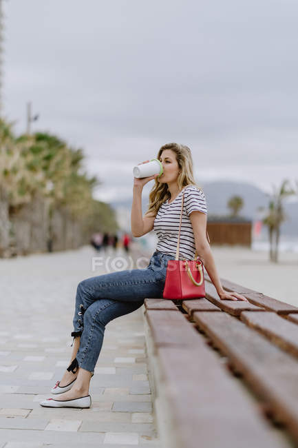 Вид сбоку женщины, пьющей из чашки кофе, сидящей на городской скамейке у моря в летний день — стоковое фото