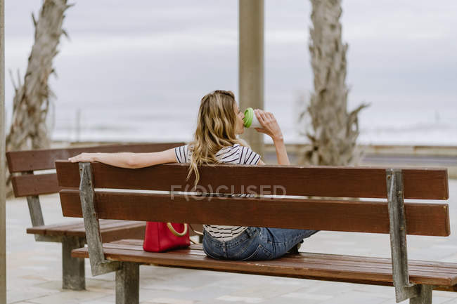 De derrière la vue de la femme joyeuse avec tasse de café à emporter assis sur le banc de la ville en bord de mer le jour de l'été — Photo de stock