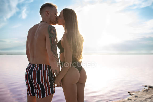 Vista trasera de pareja feliz besándose en traje de baño de pie y mirándose mientras se toman de la mano en la playa del lago rosa en Torrevieja en España en un día soleado - foto de stock