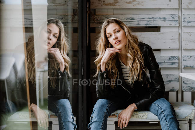 Mulher de cabelos longos na moda em jaqueta de couro e boné sentado em um banco de madeira e olhando para a câmera — Fotografia de Stock