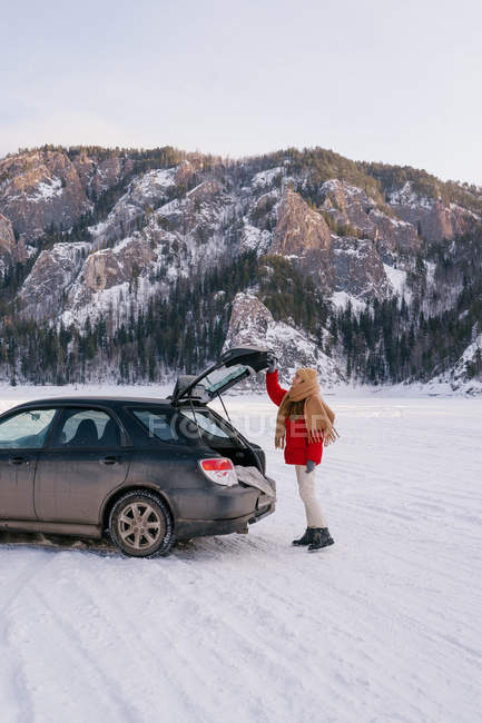 Side view of active woman in warm clothes opening car trunk while standing in snowy valley next to majestic rocks in Siberia Russia — Stock Photo