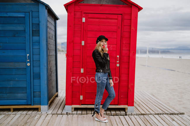 Mujer de moda en gorra negra y chaqueta de cuero comiendo una manzana roja mientras pasea por una cabaña de playa de madera - foto de stock