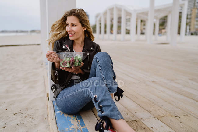 Stylische Frau in schwarzer Jacke genießt gesunden grünen Salat, während sie auf einer Holzterrasse an der Küste sitzt und weglächelt — Stockfoto