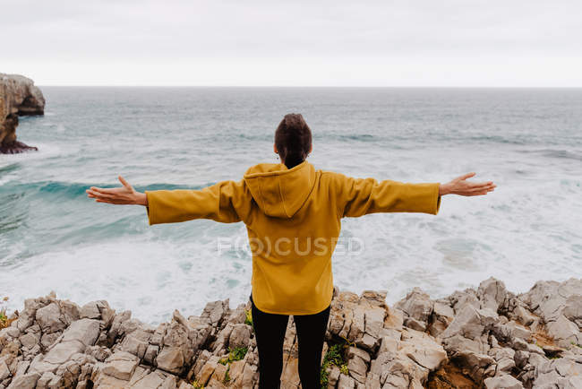 Vista trasera del viajero con capucha amarilla cálida parado solo en la orilla rocosa mirando las olas espumosas en el día nublado levantando brazos - foto de stock
