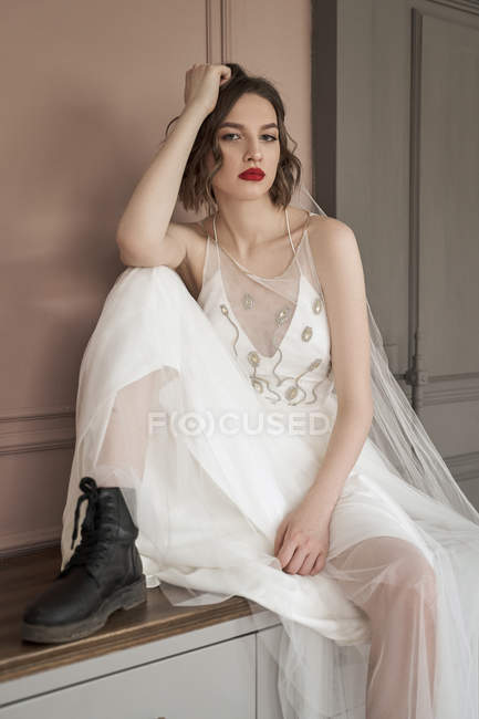 Woman with red lips in white wedding dress and black shoe leaning on hand and looking at camera against wall — Stock Photo