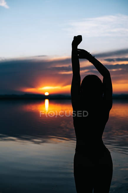Silueta de mujer de pie con los brazos levantados mientras disfruta del cielo del atardecer en la tranquila playa del lago rosa en España - foto de stock