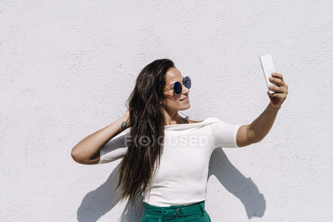 Atractiva joven hembra en traje de moda posando para selfie mientras está de pie sobre un fondo brillante - foto de stock
