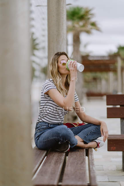 Vue latérale de femme occasionnelle joyeuse buvant du café à emporter assis sur le banc de la ville en bord de mer le jour de l'été — Photo de stock