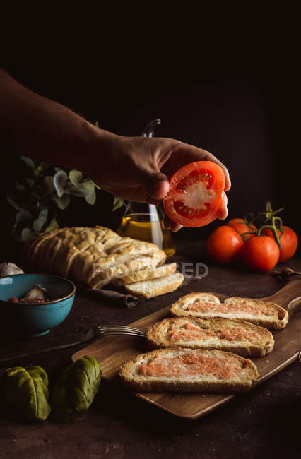 Mano de persona sosteniendo tomate sobre tostadas sobre tabla de madera - foto de stock