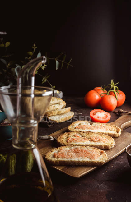 Различные специи и спелые помидоры, размещенные на доске возле кусков хлеба с соусом на столе — стоковое фото