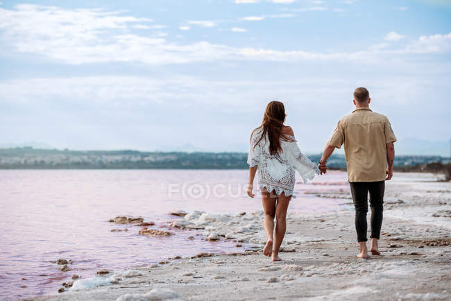 Visão traseira do casal andando em um incrível lago rosa de água do mar e céu azul de mãos dadas — Fotografia de Stock