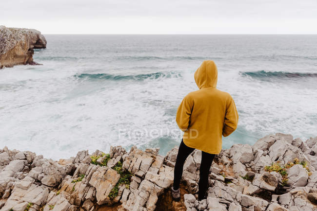 Vista trasera del viajero en sudadera con capucha amarilla cálida de pie solo en la orilla rocosa mirando las olas espumosas en el día nublado - foto de stock