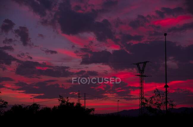 Siluetas de postes eléctricos con cables ubicados contra el cielo nublado rojo por la noche en el campo - foto de stock