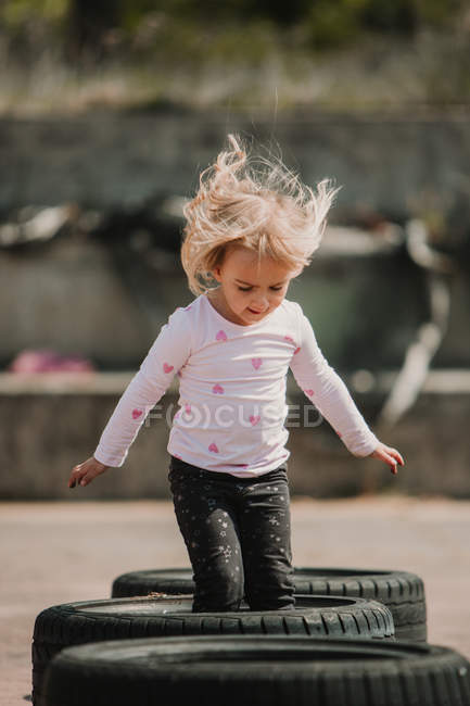 Happy joyful little girl having fun and jumping in row of black car tires while playing outdoors on summer day — Stock Photo