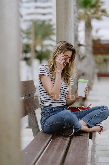 From above view of cheerful casual woman with takeaway cup of coffee sitting on city bench at seafront on summer day — Stock Photo