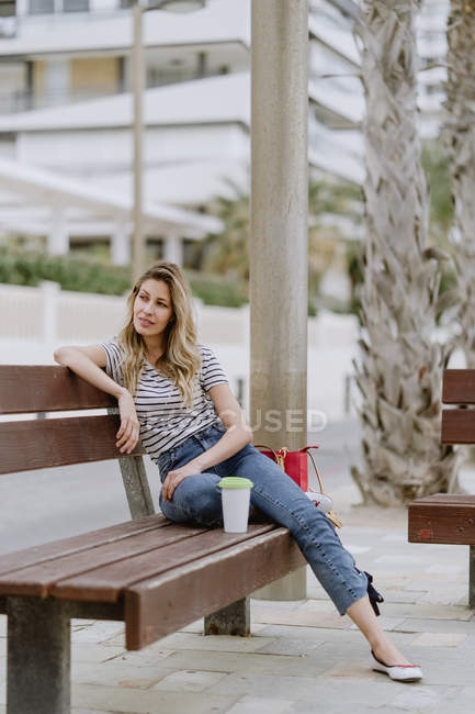 Vista laterale di allegra donna casual seduta sulla panchina della città sul lungomare il giorno d'estate guardando altrove — Foto stock