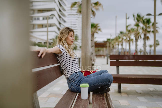 Вид сбоку жизнерадостной случайной женщины с чашкой кофе, сидящей на городской скамейке у моря в летний день — стоковое фото