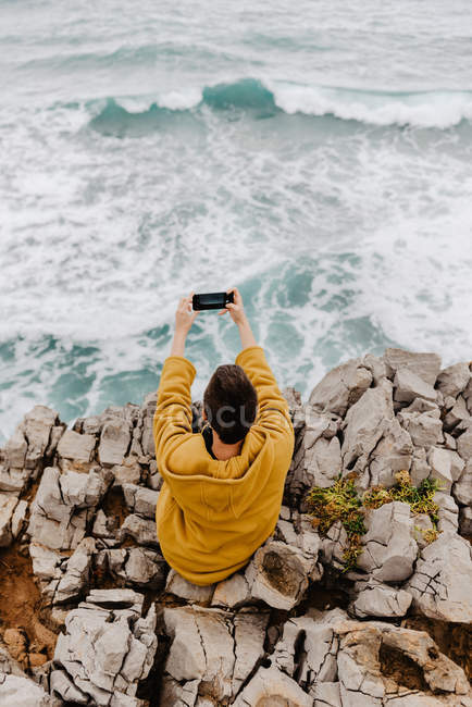 Visão traseira da mulher em camisola amarela sentada na costa rochosa e tomando selfie no telefone celular no dia nublado cinza — Fotografia de Stock