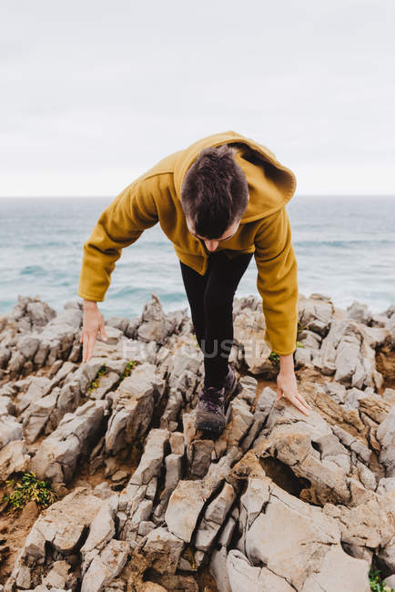 Woman in yellow warm hoodie walking alone on rocky shore with foamy waves on cloudy day — Stock Photo