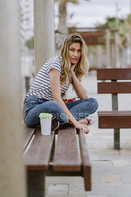 Selbstbewusste, lässige Frau, die an einem Sommertag auf einer Bank am Meer sitzt und in die Kamera blickt — Stockfoto