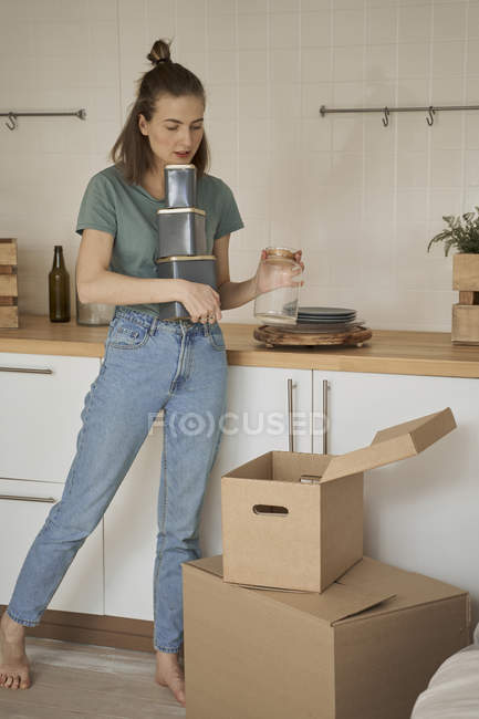 Босоногая женщина в повседневной одежде держит металлические контейнеры и вынимает стеклянную банку из коробок на кухне — стоковое фото