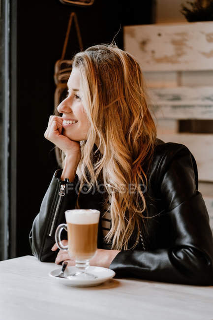 Side view of long haired trendy beautiful blonde woman sitting in a cafe shop drinking from a glass of delicious foamy coffee — Stock Photo