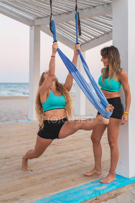 Femme prudente soutenant l'ami de fille faisant l'exercice d'acroyoga accrochant sur l'hamac sur le stand en bois à la plage — Photo de stock