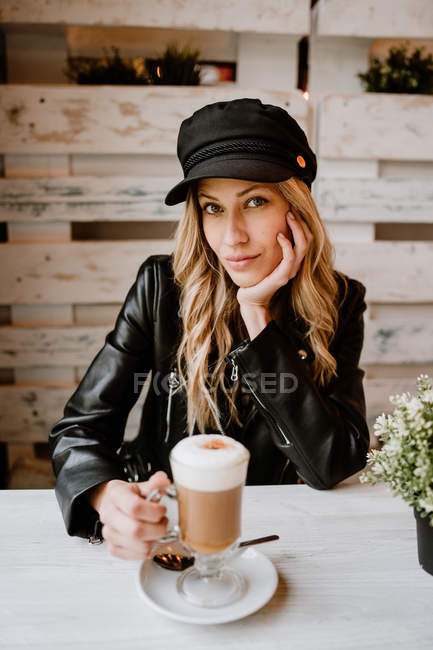 Langhaarige trendige schöne blonde Frau trinkt aus einem Glas köstlichen schäumenden Kaffee, während sie in die Kamera schaut — Stockfoto