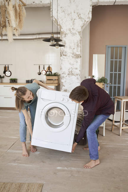 Vista lateral do homem de conteúdo descalço e mulher carregando máquina de lavar roupa branca enquanto se move para a nova casa — Fotografia de Stock