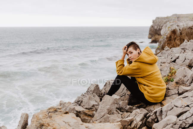 Vista lateral de la mujer en sudadera con capucha cálida amarilla sentada sola en olas rocosas en el día nublado y mirando a la cámara - foto de stock