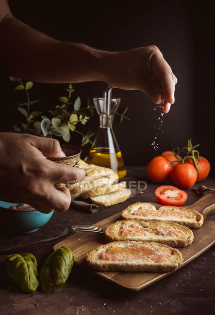 Persona irreconocible derramando sal de trozos de pan fresco con salsa y aceite mientras cocina tostadas sobre fondo negro - foto de stock