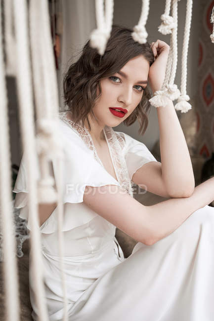 Gorgeous woman with red lips in white dress looking at camera while sitting on floor beside sofa — Stock Photo