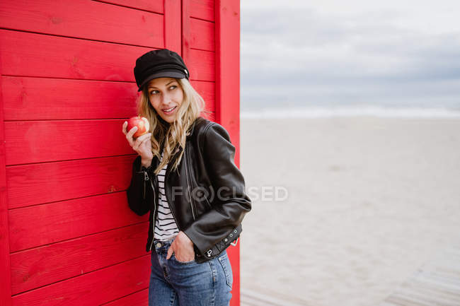 Donna in berretto nero e giacca di pelle che mangia una mela rossa mentre si appoggia su cabine da spiaggia in legno — Foto stock