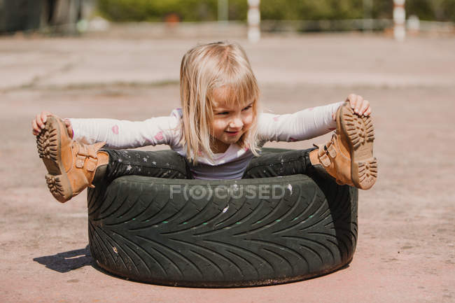Feliz adorável menina sentada dentro pneu do carro enquanto se diverte e jogando ao ar livre no dia de verão — Fotografia de Stock