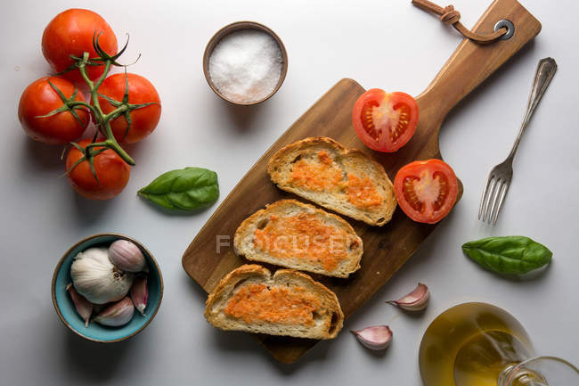 Varie spezie e pomodori maturi posti sul tagliere vicino a pezzi di pane con salsa su fondo bianco — Foto stock