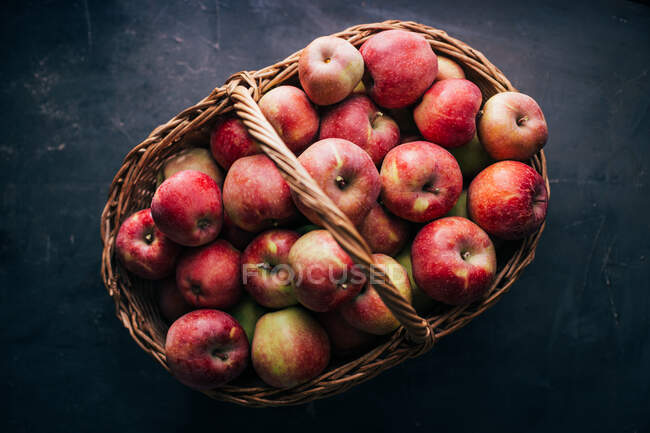 Manzanas rojas frescas sobre mesa oscura y en canasta de mimbre sobre fondo oscuro - foto de stock