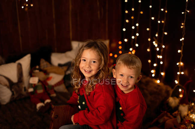 Приваблива маленька дівчинка і хлопчик сидять на дерев'яному коні гойдалці в кімнаті повний різдвяних прикрас і дивляться в камеру — стокове фото