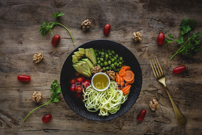 Macarrão vegan cru de abobrinha com ervilhas, tomates cereja, abacate, cenouras, nozes e azeite em tigela servida em fundo de madeira — Fotografia de Stock