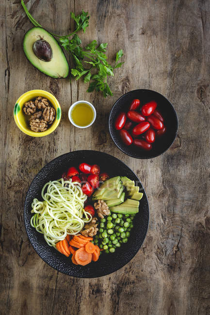 Zucchini rohe vegane Pasta mit Erbsen, Kirschtomaten, Avocado, Karotten, Nüssen und Olivenöl in einer Schüssel auf Holzboden serviert — Stockfoto