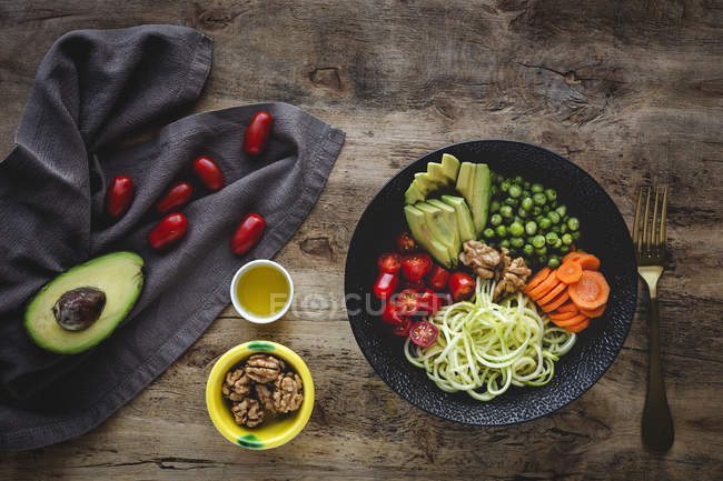 Macarrão vegan cru de abobrinha com ervilhas, tomates cereja, abacate, cenouras, nozes e azeite em tigela servida em fundo de madeira — Fotografia de Stock