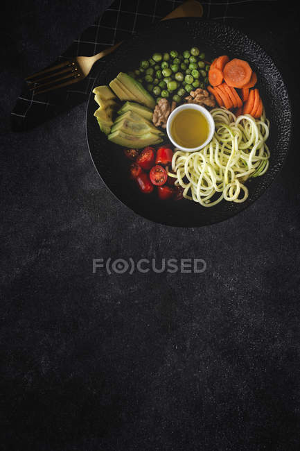 Macarrão vegan cru de abobrinha com ervilhas, tomates cereja, abacate, cenouras, nozes e azeite em tigela servida em fundo escuro — Fotografia de Stock