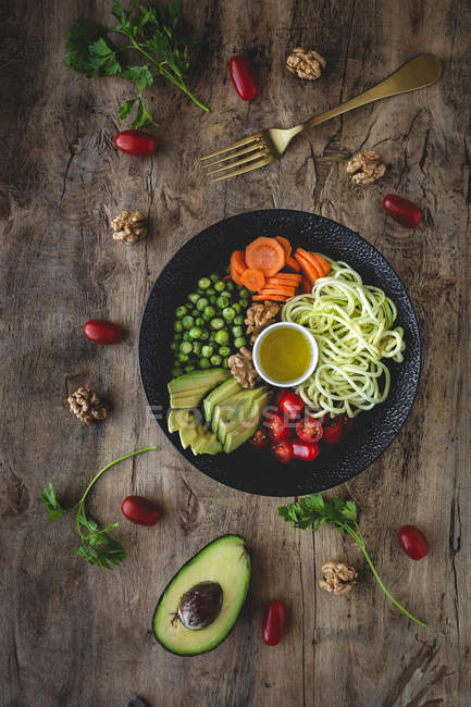 Zucchine crude vegane con piselli, pomodorini, avocado, carote, noci e olio d'oliva in ciotola servite su fondo di legno — Foto stock