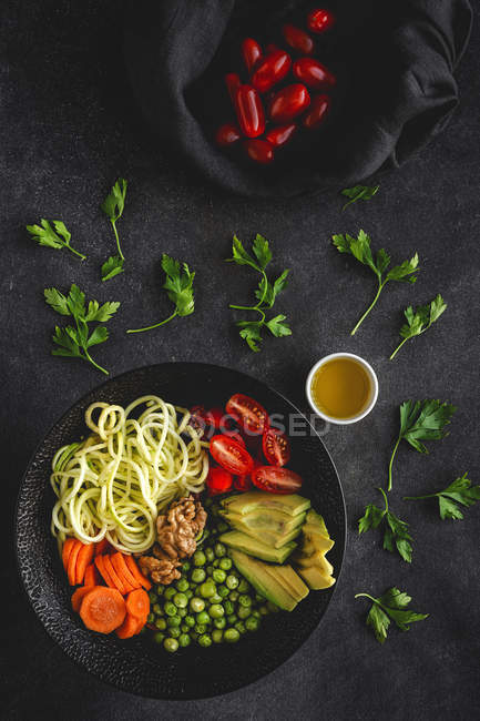 Zucchini rohe vegane Pasta mit Erbsen, Kirschtomaten, Avocado, Karotten, Nüssen und Olivenöl in einer Schüssel auf dunklem Hintergrund serviert — Stockfoto