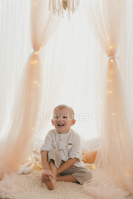 Tender little boy sitting, smiling and looking away with fairy lights and stylish draperies — Stock Photo