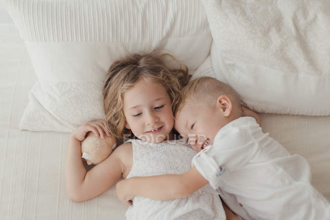 From above of happy male and female kids in white clothing lying on bed in embrace and smiling — Stock Photo