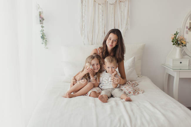 Positive junge Brünette sitzt auf dem Bett und umarmt kleine Kinder, während sie in die Kamera auf stilvolles Schlafzimmer blickt — Stockfoto