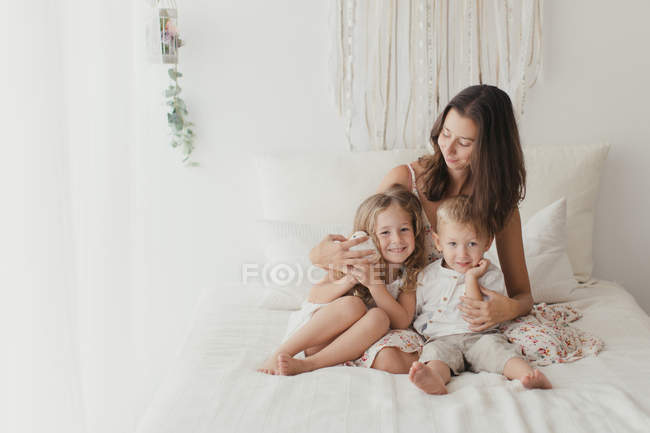 Позитивная юная брюнетка сидит на кровати и обнимает маленьких мальчиков и девочек в стильной спальне — стоковое фото